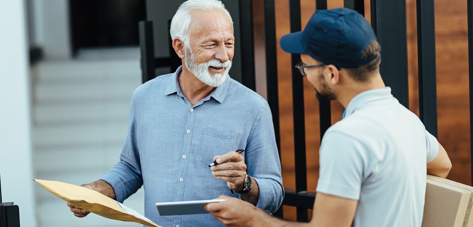 Ein älterer Mann hält einen Umschlag und spricht mit einem jüngeren Boten, der ein Paket und ein Tablet in der Hand hat. Beide lächeln.